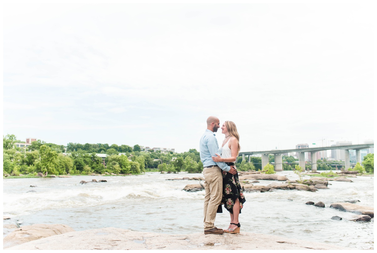 couple standing in front of james river backdrop with sand and rocks in background sunny day in the summer richmond virginia engagement photos and inspo by sarah & dave photography virginia washington dc and annapolis and charlottesville wedding elopement destination and engagement photographer