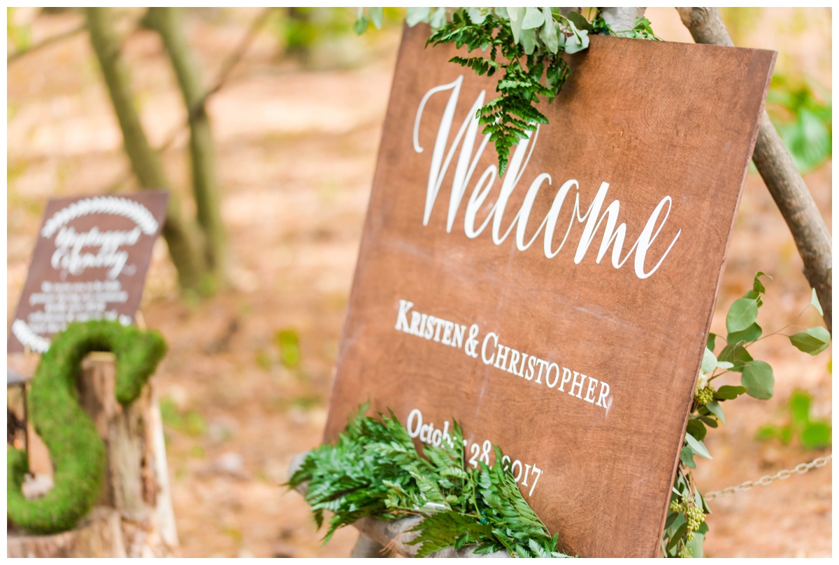 Whimsical Woodland Fall Wedding at mountain memories at thorpewood in thurmont maryland in october by sarah & dave photography richmond wedding photographer wooden forest inspired welcome sign with white letters greenery ferns and log easel outdoor ceremony in the woods