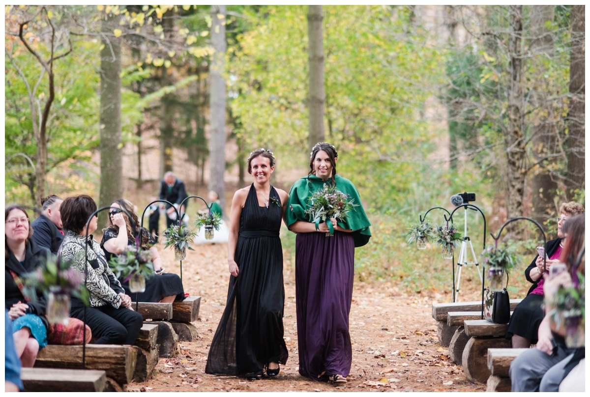Whimsical Woodland Fall Wedding at mountain memories at thorpewood in thurmont maryland in october by sarah & dave photography richmond wedding photographer forest inspired outdoor ceremony in the woods bridesmaid and groomwoman walking down aisle log tree stump seating seats black dress deep dark purple bridesmaid dress with green cape bridesmaid cape fall wedding attire dress inspiration
