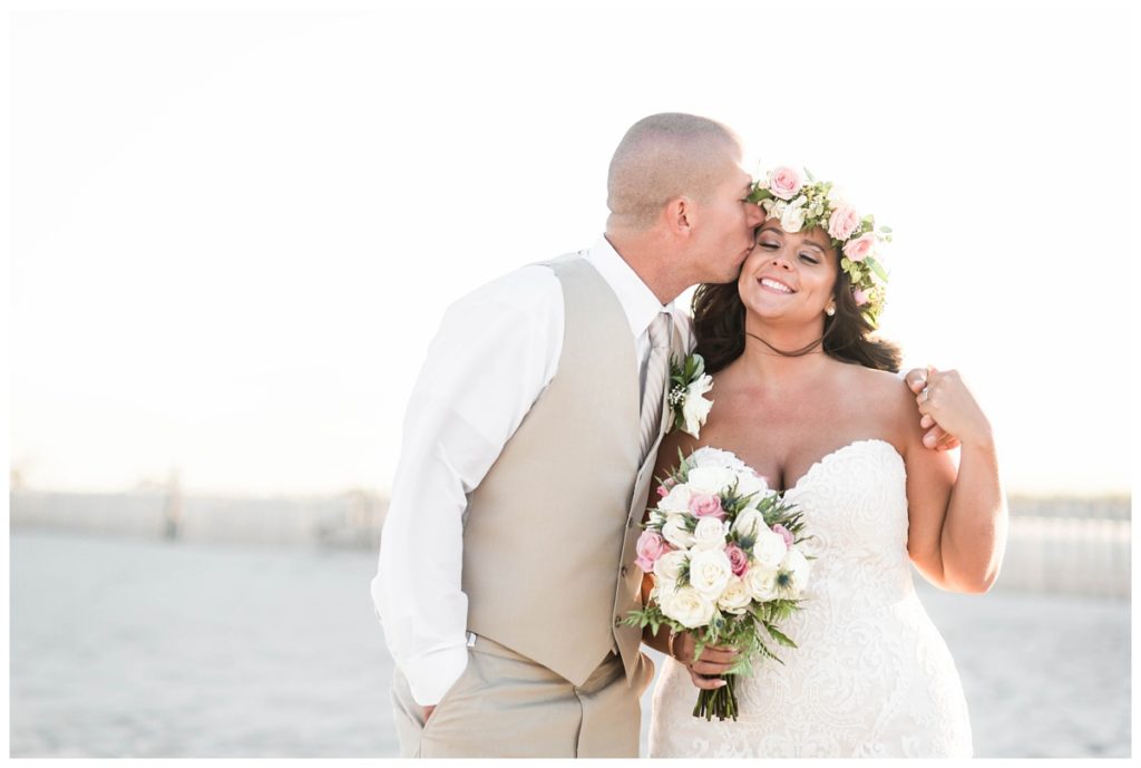 pink and gold summer beach wedding in september couple kissing on sand bride is wearing flower crown and holding bouquet smiling groom is wearing light tan suit perfect sunset photo