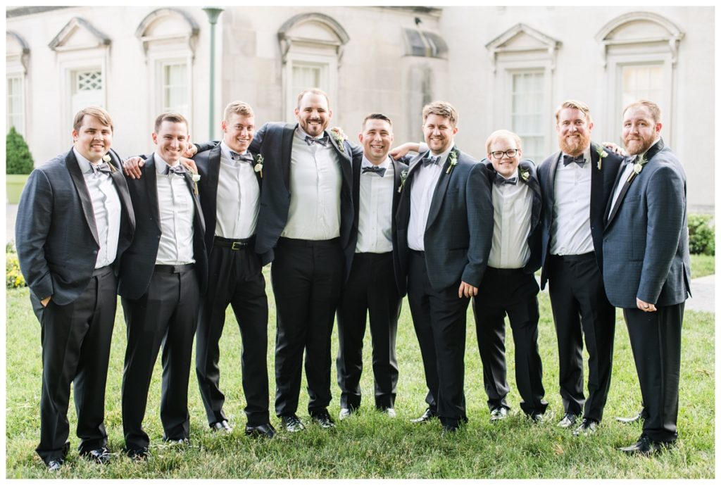 groom and groomsmen photo outdoors in the summer wearing dark suits and bowties laughing