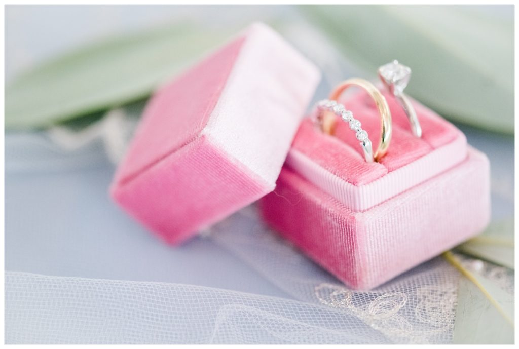 pink ring box flatlay and wedding rings against bridal veil and a few green sprigs of leaves and light blue backdrop