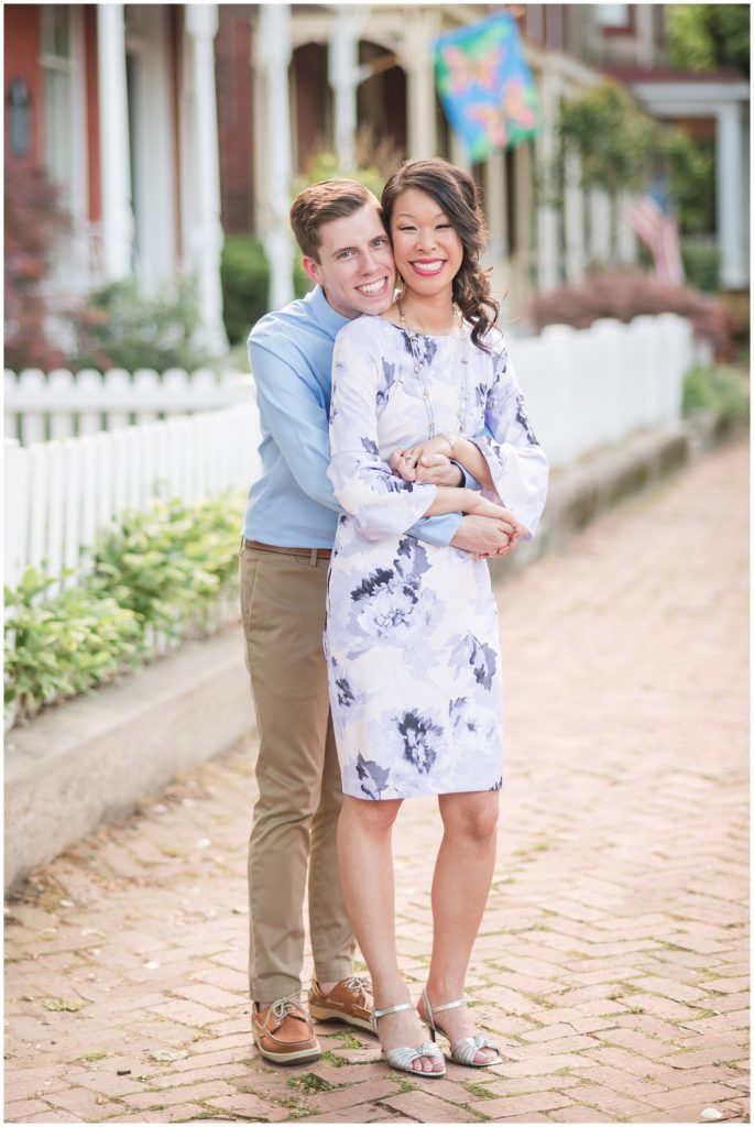 Engagement photos near Libby Hill Park in Church Hill neighborhood of Richmond Virginia RVA - spring - springtime - smiling couple outdoors in front of brick rowhomes townhomes homes buildings in front of white picket fence and flowers