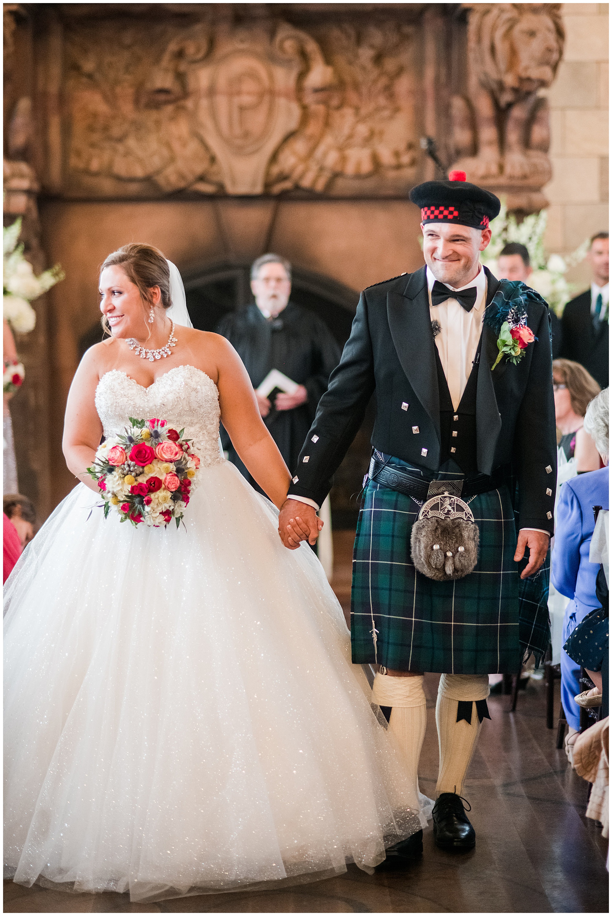 just married photo. bride and groom holding hands and walking down the aisle. groom is wearing kilt and tartan. bride is wearing princess cut ballgown wedding dress that is sparkly. dover hall estate ballroom venue is in the backdrop.